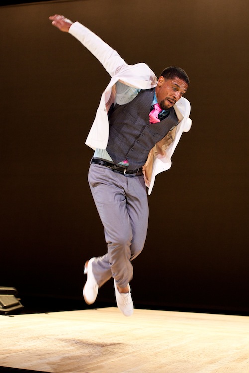 Tapper Jason Samuels Smith in a white blazer and colorful tip leaps into the air. His white tap shoes are off the ground.
